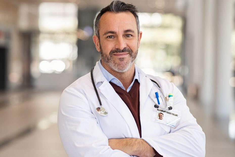 Médico em hospital com jaleco e estetoscópio, de braços cruzados, transmitindo confiança em ambiente clínico.