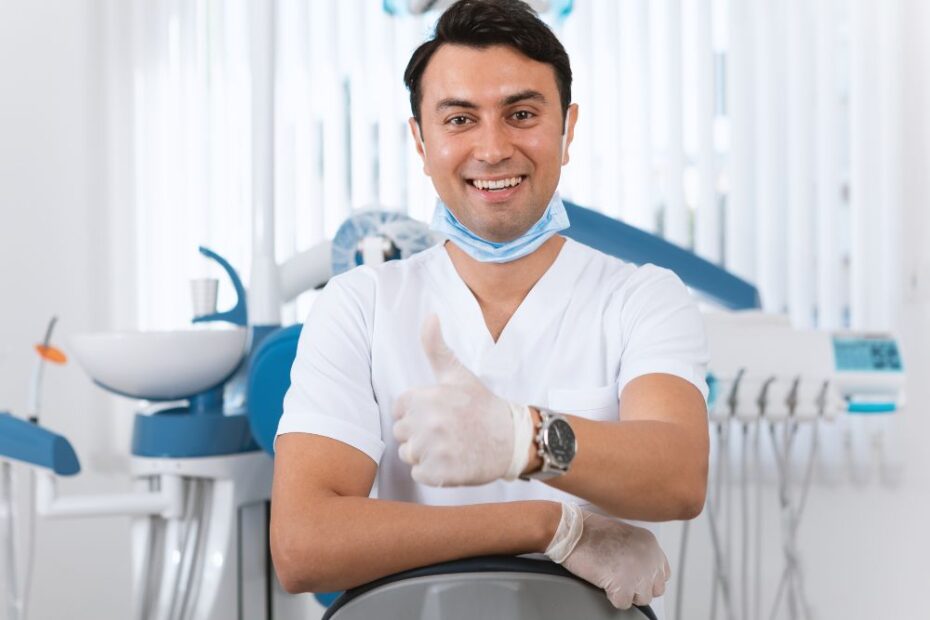 Dentista em consultório odontológico sorrindo e fazendo sinal positivo com o polegar, com equipamentos ao fundo.